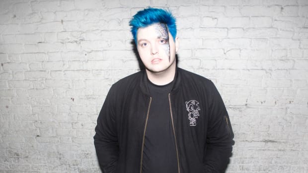 A color press photo of English DJ/producer Flux Pavilion (real name Joshua Steele) standing in front of a white brick wall.