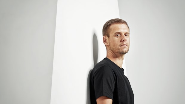 A color press photo of Armin van Buuren wearing a black tee shirt and standing against a giant letter A.