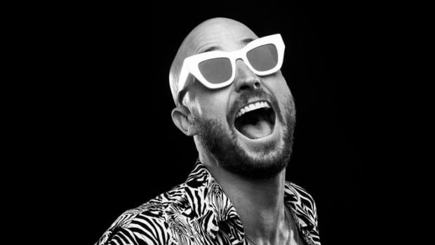 A black-and-white photo of Australian DJ/producer FISHER wearing white sunglasses and a zebra-print jacket over a black background.