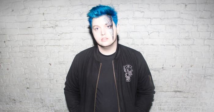 A color press photo of English DJ/producer Flux Pavilion (real name Joshua Steele) standing in front of a white brick wall.