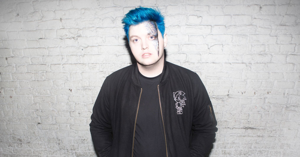 A color press photo of English DJ/producer Flux Pavilion (real name Joshua Steele) standing in front of a white brick wall.