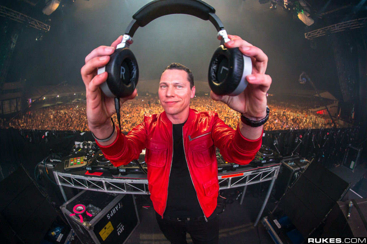Tiesto holding headphones and wearing a red jacket while DJing in front of a big crowd.