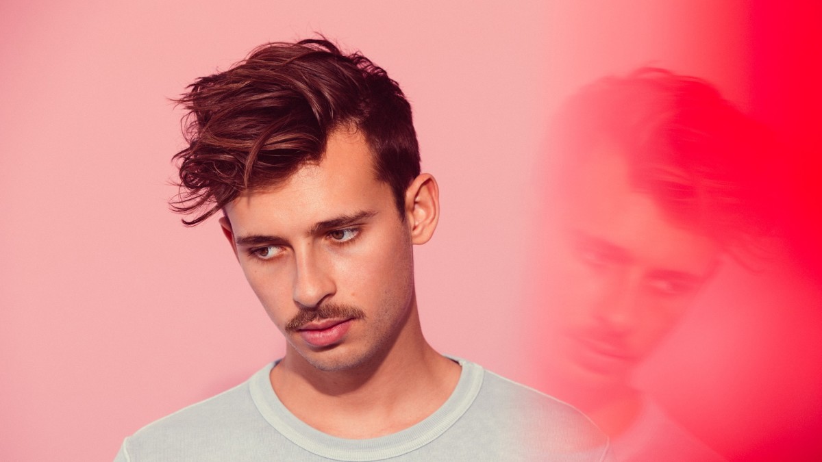 A photo of Australian DJ/producer Flume (real name Harley Edward Streten) in front of a reddish-peach background with a hazy reflection of his face.