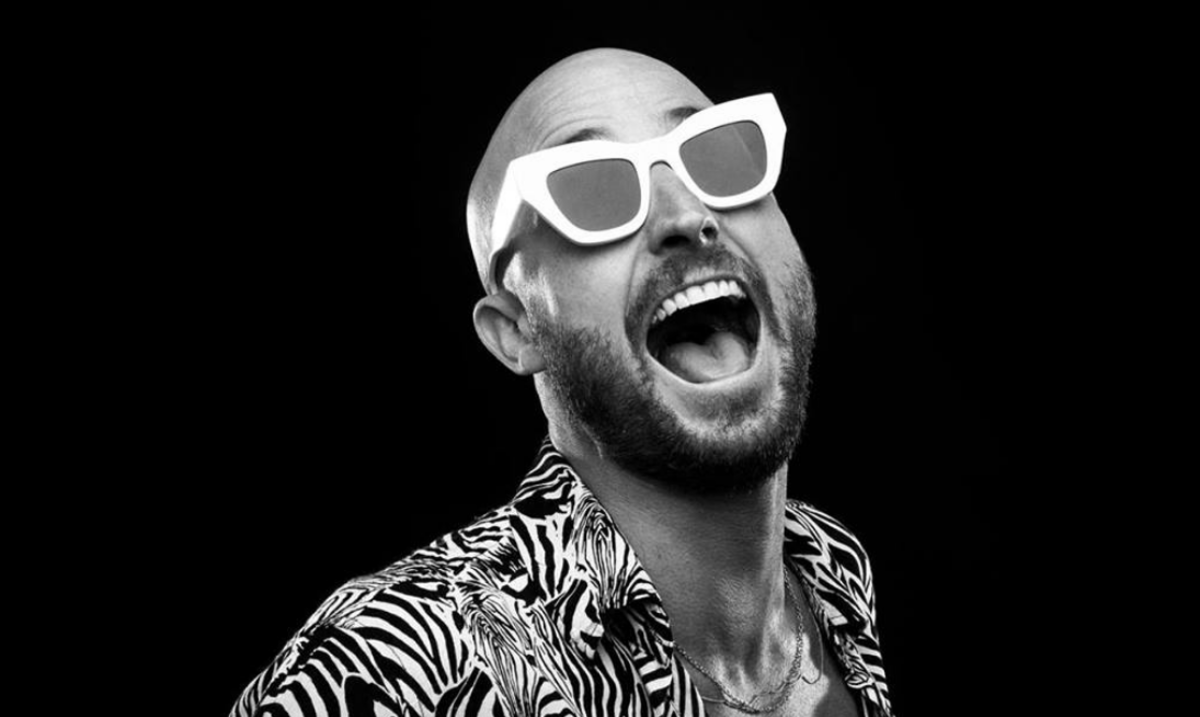 A black-and-white photo of Australian DJ/producer FISHER wearing white sunglasses and a zebra-print jacket over a black background.